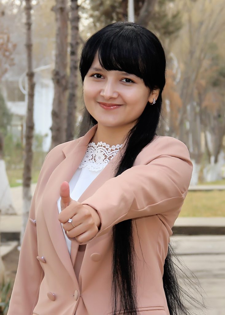 Young Central Asian woman standing outside in front of trees and a house. She's got black hair and bangs and earrings and is in a lacy white top and a pink jacket and is giving a thumbs up. 