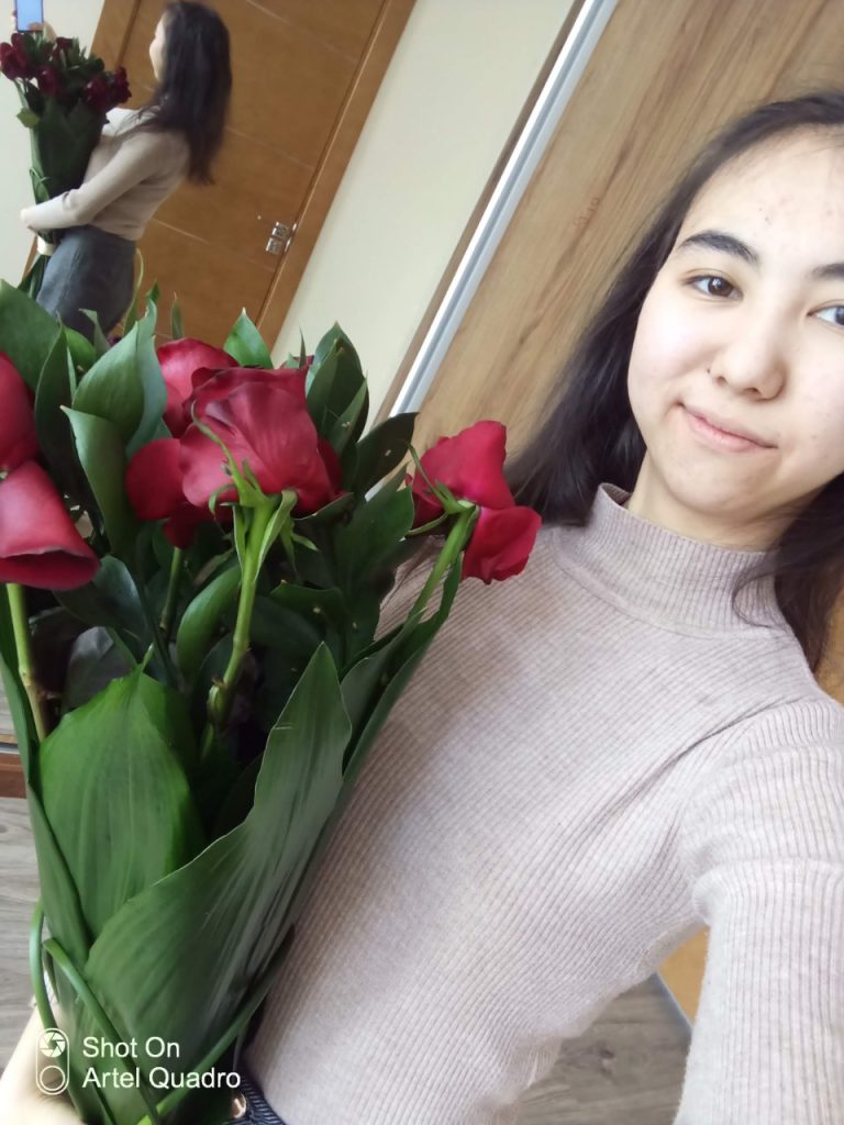 Young Central Asian teen girl with long dark hair and a beige sweater holding a bouquet of roses. She's inside standing in front of a mirror. 