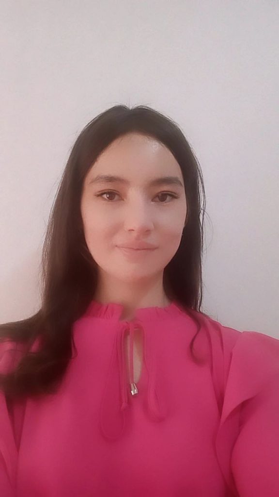 Young Central Asian woman with dark hair and brown eyes standing up, wearing a pink sweater.