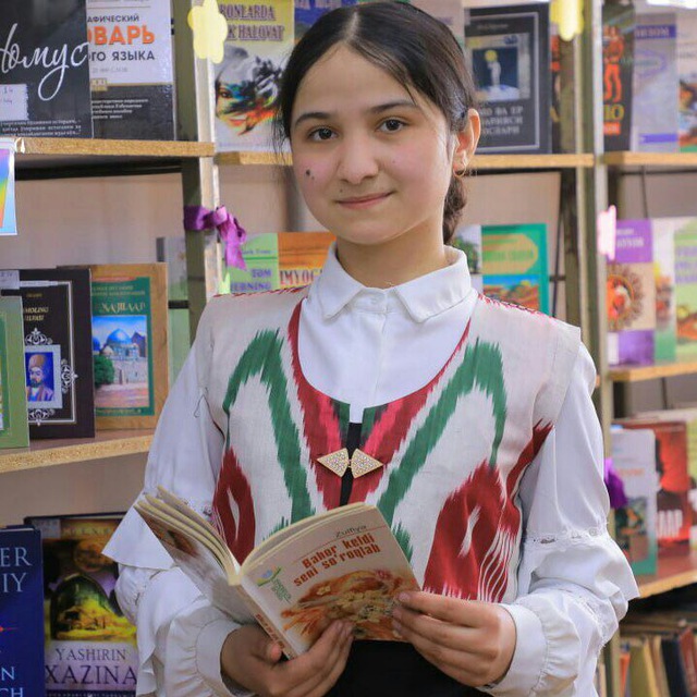 Young Central Asian girl with long dark hair behind her head and a white collared shirt with a red and green vest. She's standing in front of a bookshelf reading a book. 