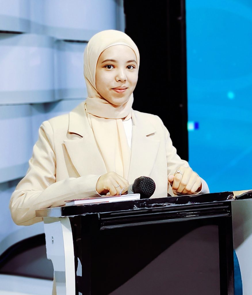 Central Asian woman with a beige headscarf and professional jacket standing at a podium with a microphone.