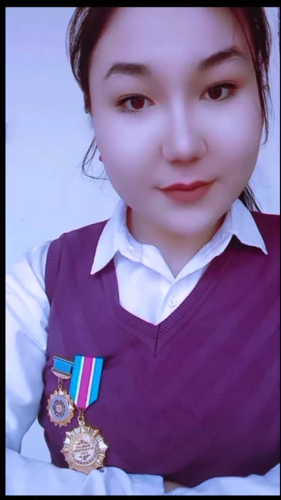 Closeup headshot of a young Central Asian woman with black hair and black eyes and a purple sweater over her collared shirt. She's wearing medals on her breast. 