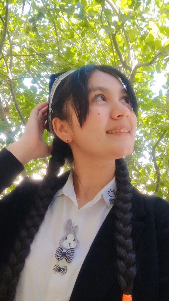 Teenage Central Asian girl with black hair and eyes and a white headband and black braids and a kitty on her white polo shirt and a black jacket. She's standing outside under a few trees on a sunny day. 