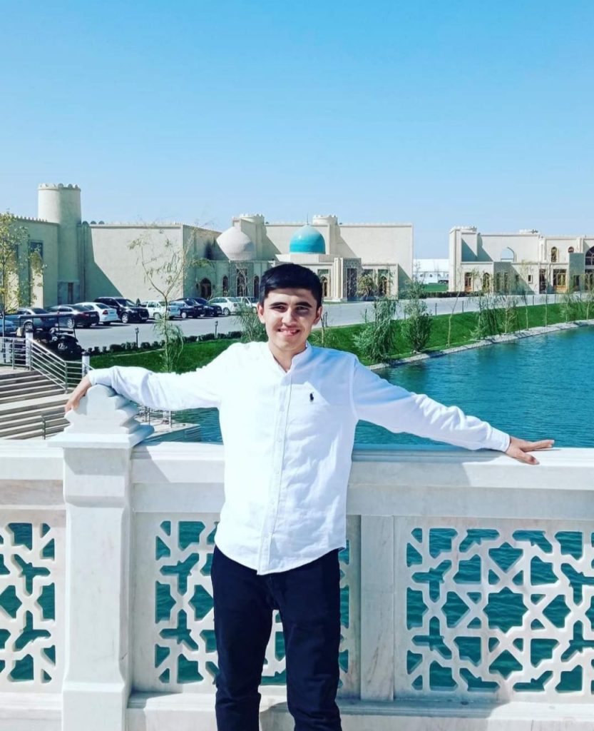 Young Central Asian teen guy in a white collared shirt and black pants standing in front of a river or late and trees and buildings off of a bridge. 