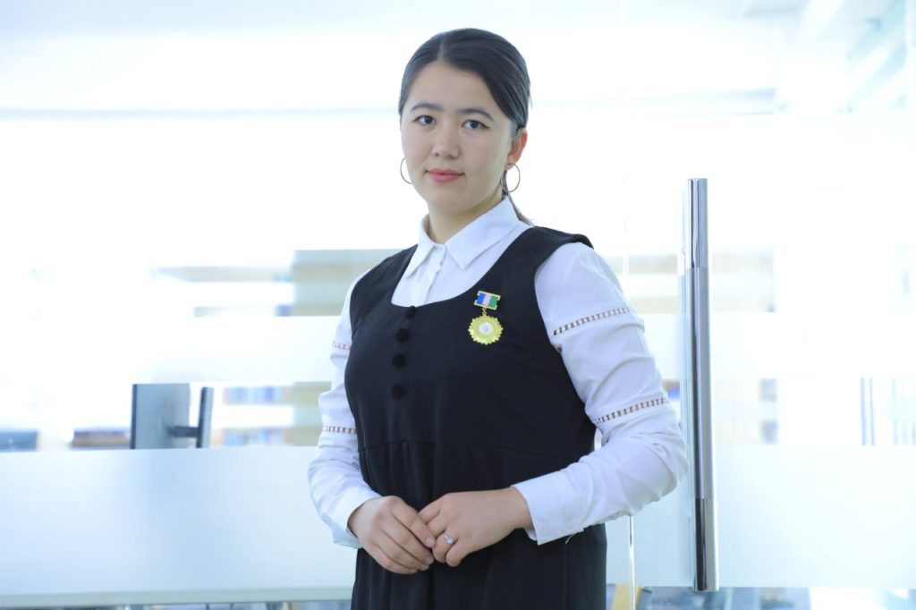 Central Asian woman standing in front of a nondescript office background. She's got dark hair, earrings, a white collared shirt, and black overalls with a medal on her right breast. 