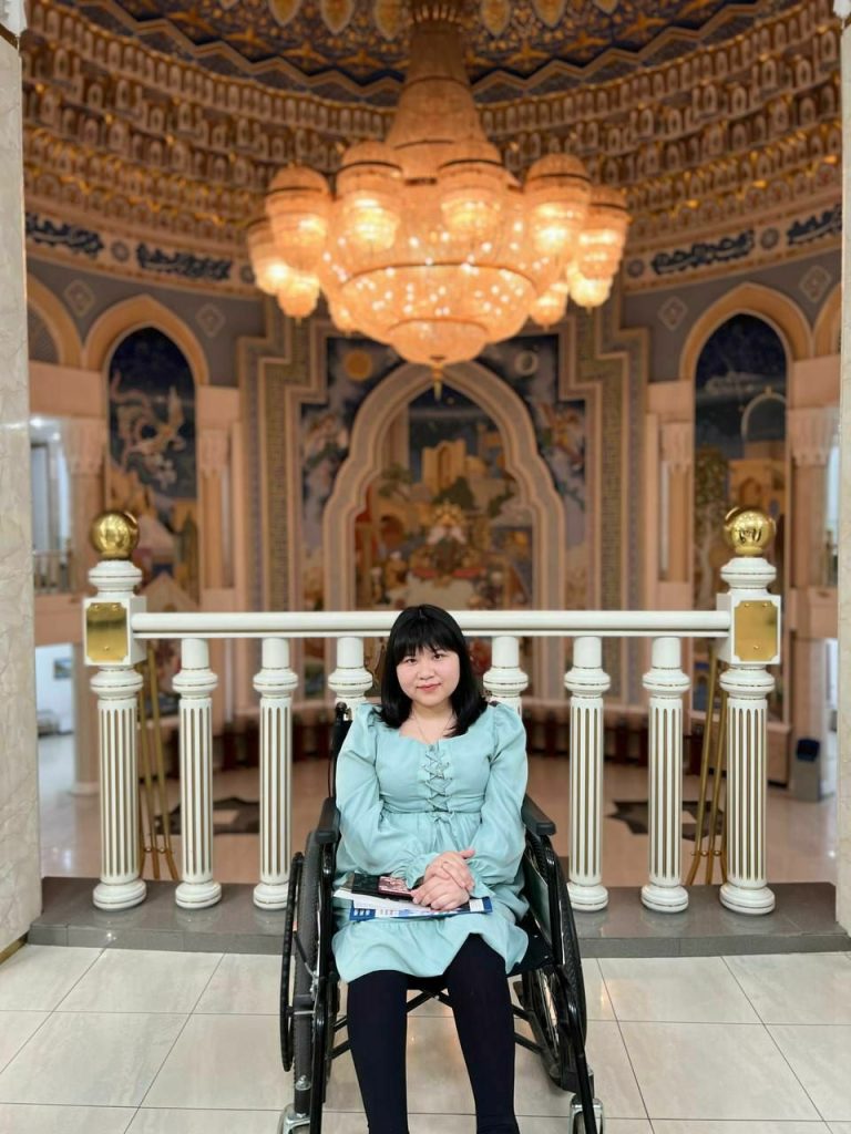 Young Central Asian woman in a short green dress and black stockings, in a wheelchair. She's posing in front of a bannister and painted walls and a chandelier. 