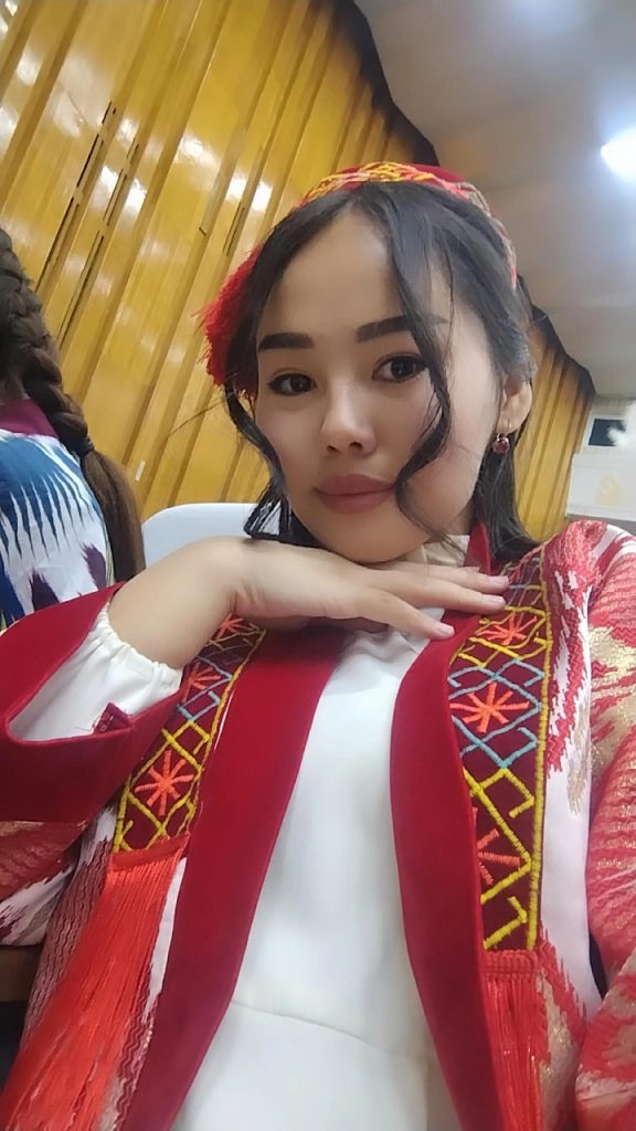 Young Central Asian woman with dark, short, slightly curly hair, a red and yellow and pink headscarf, a white blouse and a red patterned vest. 