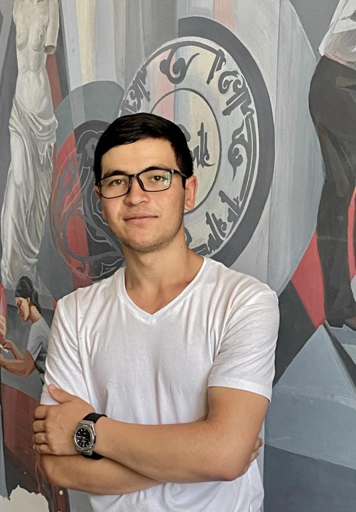 Central Asian young man with dark short hair, glasses, and a white tee shirt and wristwatch. He's in front of a patriotic painting of something to do with Uzbek history.