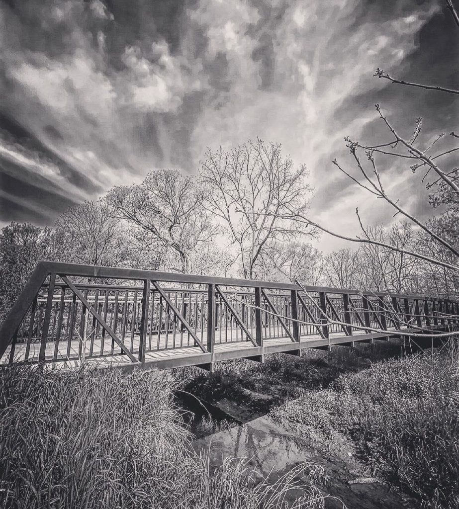 Black and white photo of a wood bridge with trees and clouds in the background.
