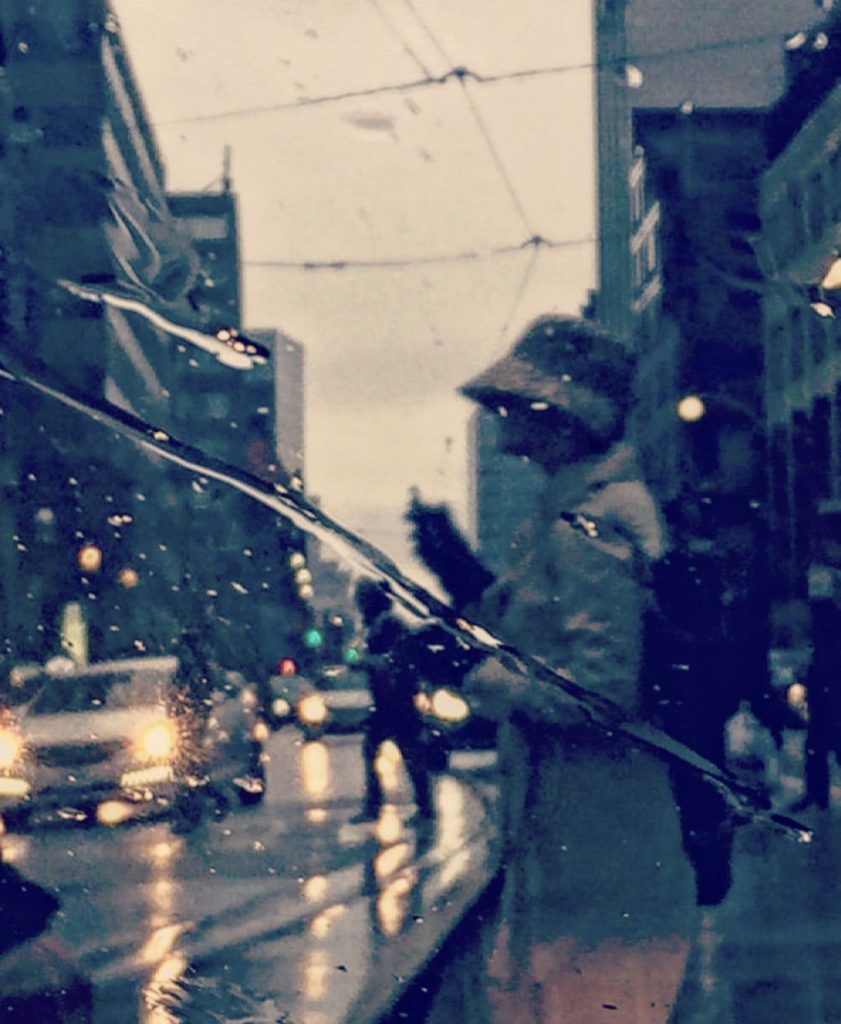 Young person of color in a hat and coat standing by the side of a city street in the rain. Cars are driving by and people are walking. 