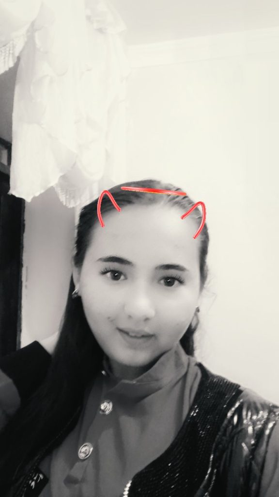 Teen Central Asian girl with cat ears drawn on her head in a Snapchat filter. She's in black and white with a collared shirt and jacket. 