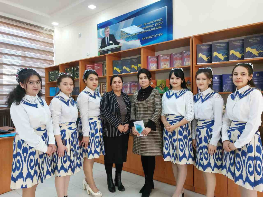Two women teachers in a short dress and in a skirt and jacket stand in a library flanked by young teen girls. The girls are in school uniforms that are collared white shirts with blue designs on the collar and white skirts with blue designs. 