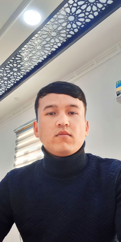 Young Central Asian man stands in a classroom with a bookshelf to his right and a patterned divider above him. He's got a serious face and a black sweater.