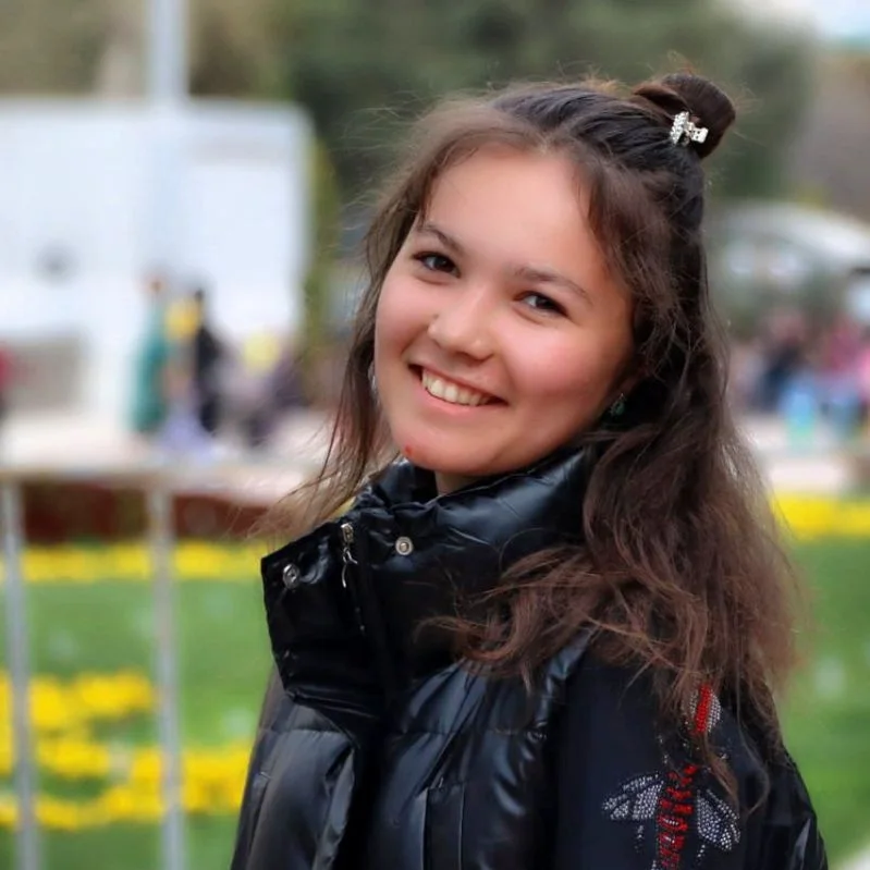 Smiling young adult Central Asian woman, hair up in a bun and a puffy black coat. She's outside on a green lawn. 