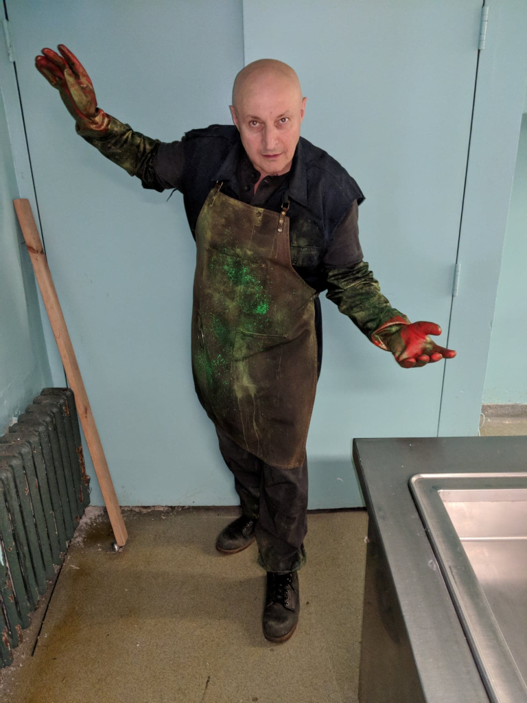 White middle aged bald man with brown overalls and green and yellow paint on his clothes. He's standing in a room near a white wall and a radiator.