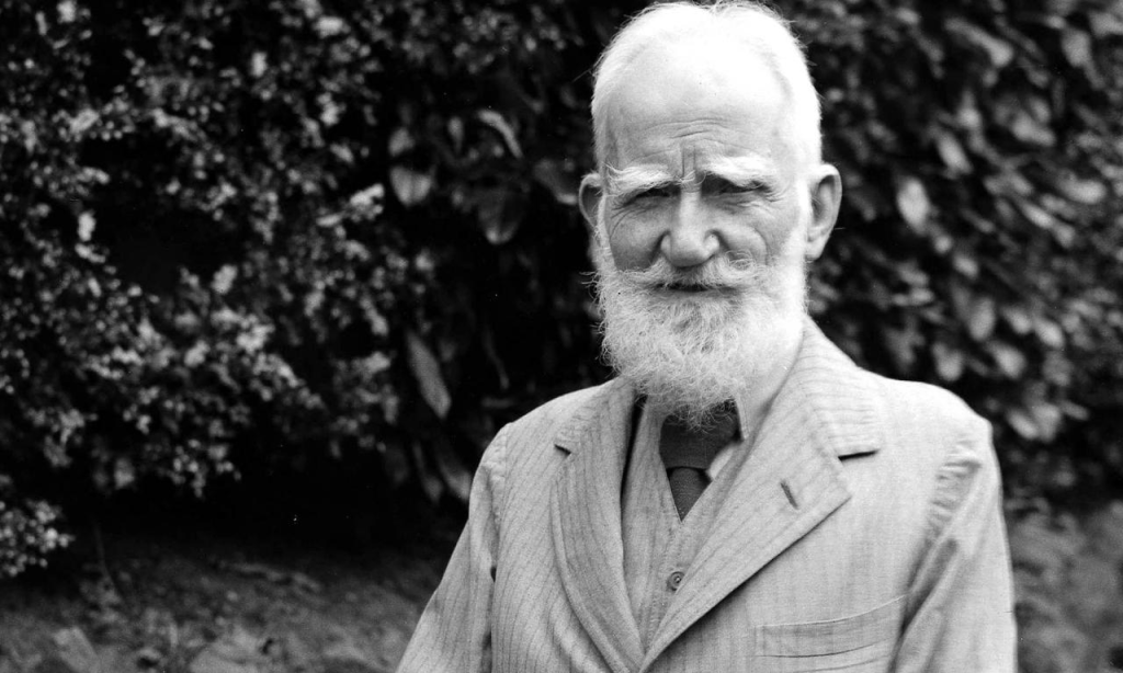 George Bernard Shaw, old man with thinning white hair and a beard and coat, in black and white outside on a lawn in front of a bush. 