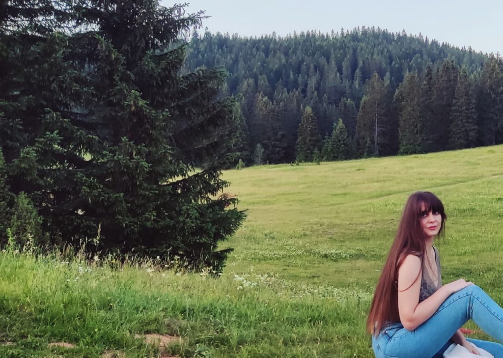 Young white woman with long dark hair sitting in a meadow clearing in a forest. She's got a green top and blue jeans. 