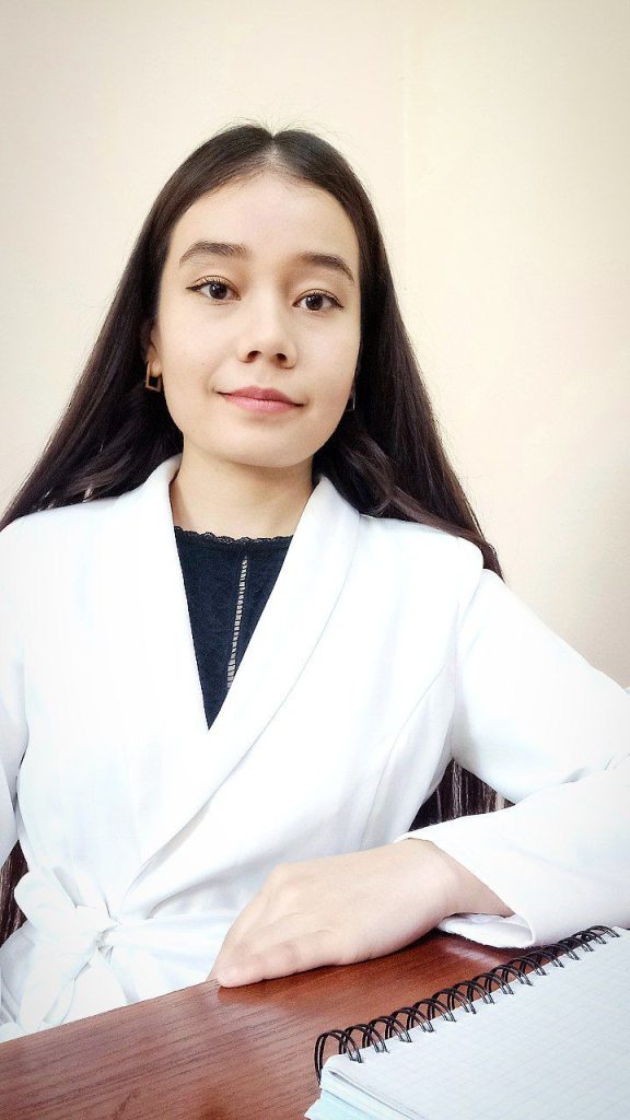 Central Asian teen girl with long straight dark hair, brown eyes, a white collared jacket, and a lacy black blouse. She's sitting at a desk with her hand on the wood and a notebook next to her. 