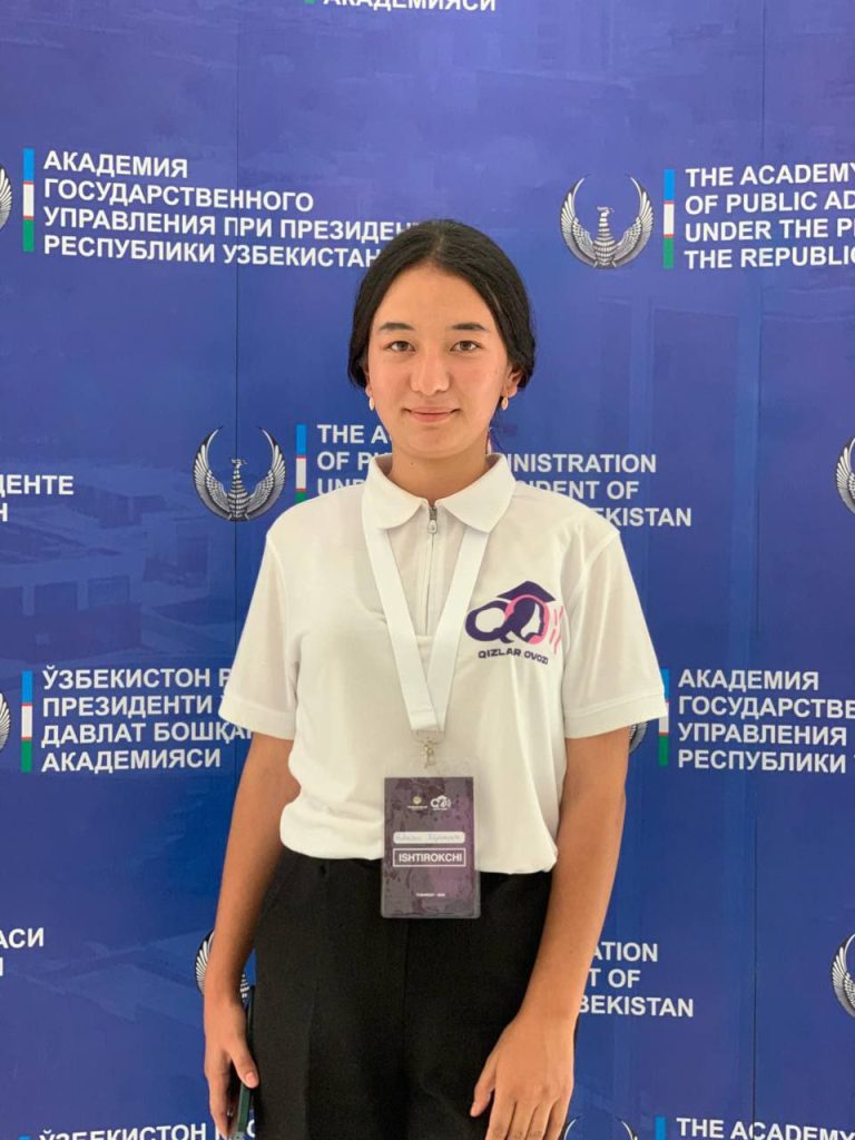 Central Asian teen girl with short dark hair and a white collared school uniform jacket stands in front of a purple banner.