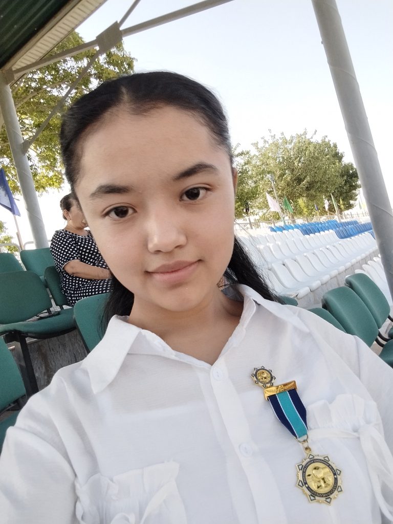 Central Asian teen girl with long hair in a braid and a white collared shirt school uniform sitting outside where there are a group of chairs and a few trees. 