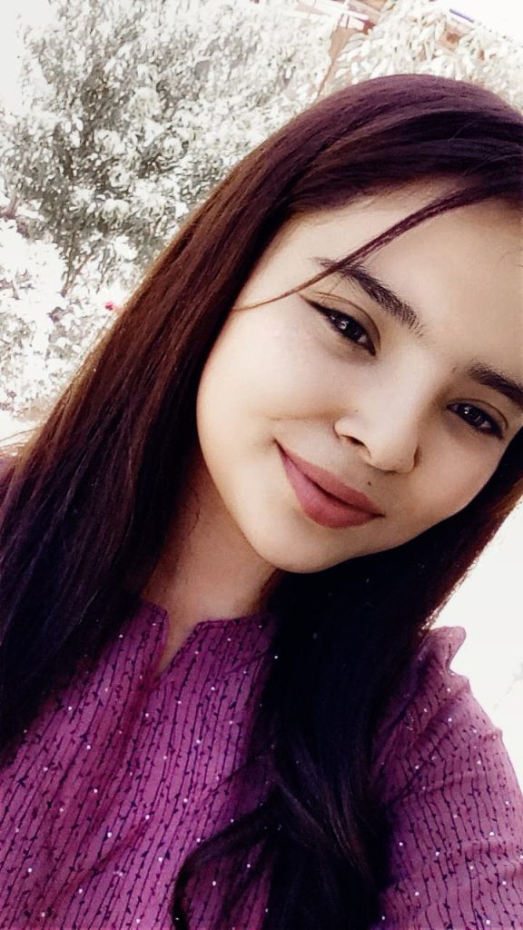 Young Central Asian woman posing at a diagonal angle. She's got slightly red/purple hair and a sparkly purple and black blouse. 