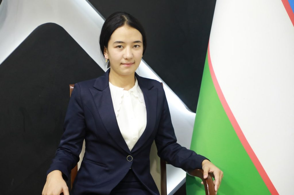 Young Central Asian teen girl with dark straight hair and a black coat and a white collared blouse standing in front of the Uzbek flag. 
