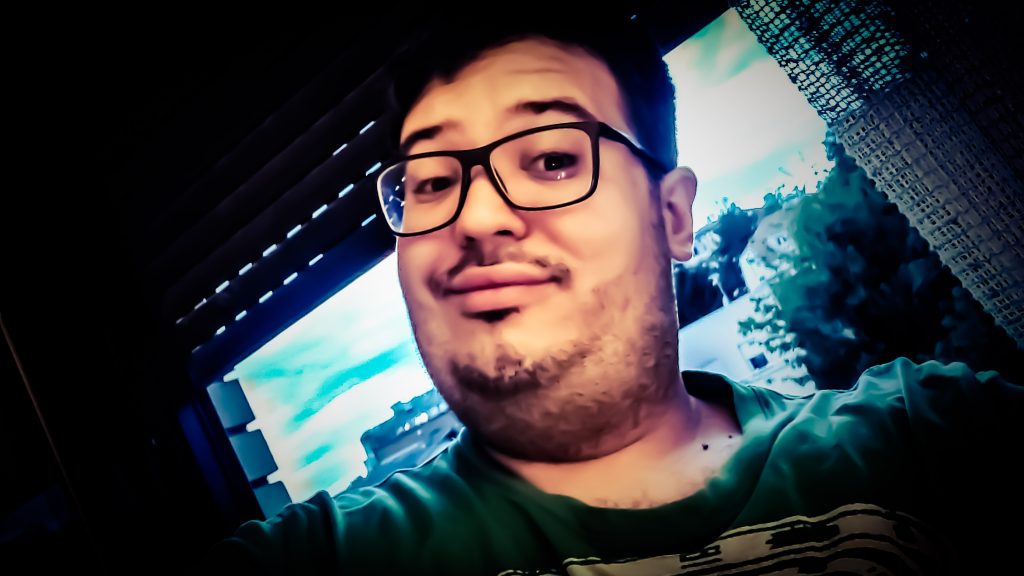 Young adult white man with a green tee shirt and reading glasses on. He'/s standing in a room in front of an open window. 