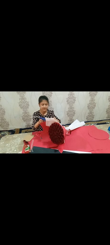 Young Central Asian woman with long straight dark hair behind her head and a black and white blouse. Shei's at a table making some sort of craft out of red paper. 