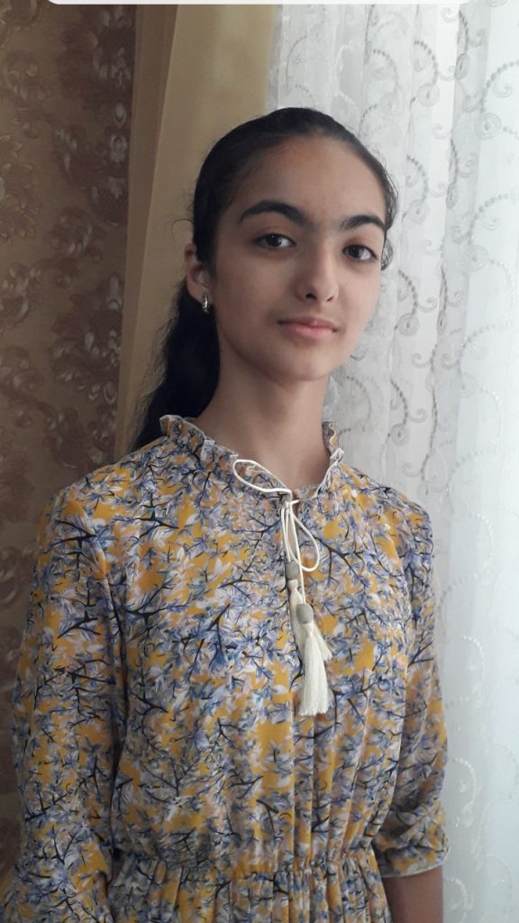 Young Central Asian teen girl with long dark hair tied behind her back. She's standing in front of a lace curtain and in a blue and yellow and white dress. 