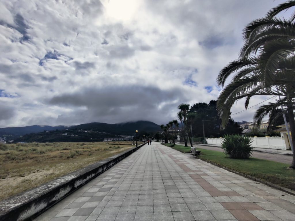 Looming dark clouds ahead on a concrete path with a grassy field to one side and a watered lawn and palm trees on the other. Hills in the distance. 
