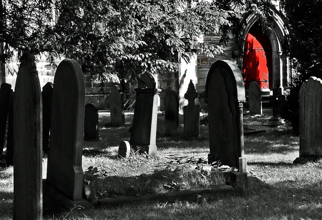 Black and white cemetery with tall rounded headstones lined up in the grass under a tree. One chapel or tomb has a red door. 