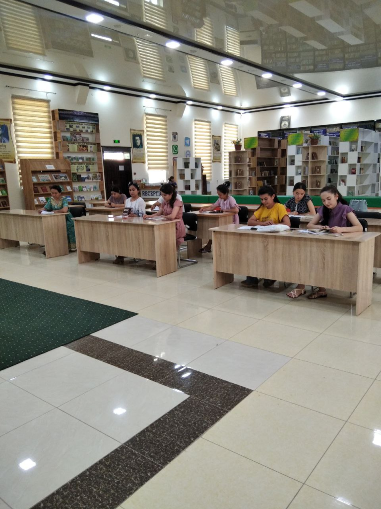 Elementary or middle grade Central Asian students seated at desks with books and paper in a library with books and magazines on racks. 