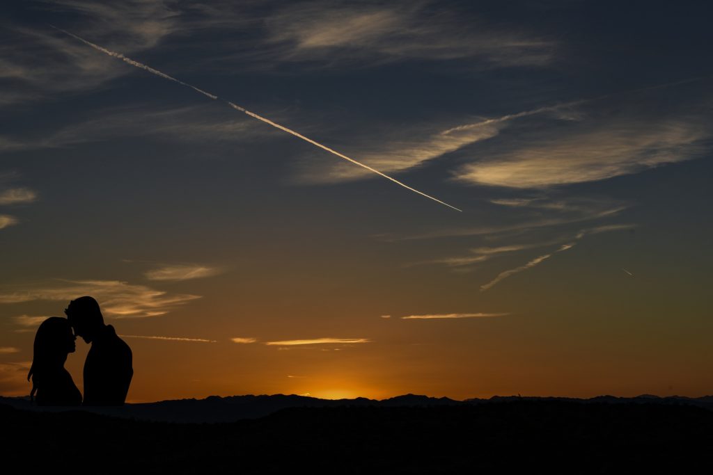 Yellow sunset behind the horizon, wispy white clouds in the sky, blue above the sunset. Couple embraces in the lower left corner. 