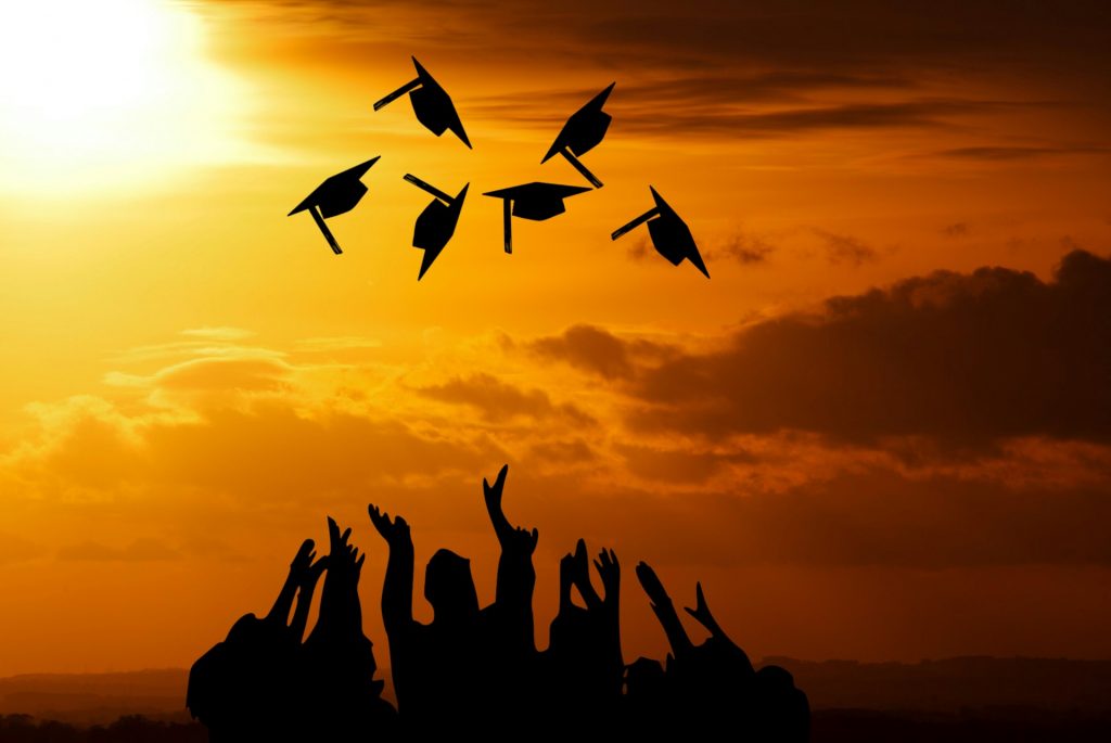 Silhouette of a sunset with graduates throwing their caps in the air against the orange cloudy sky.