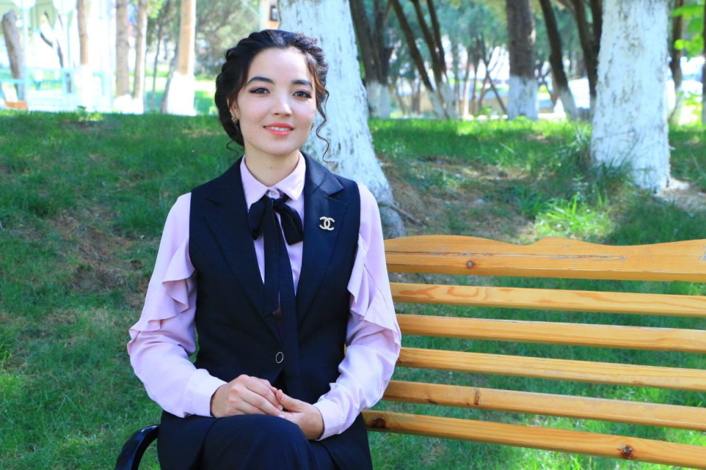 Young Central Asian woman with dark curly hair, brown eyes makeup, a pink ruffly blouse, and a black best. She's in front of trees, grass, and a wooden bench.