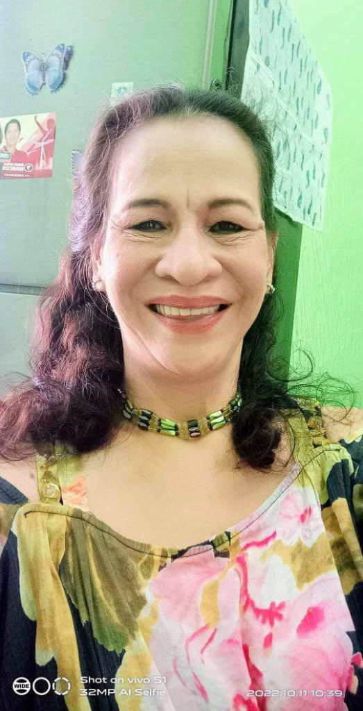 Light skinned Filipina woman with reddish hair, a green and yellow necklace, and a floral pink and yellow and green blouse.