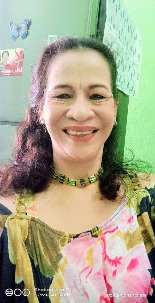 Older light skinned Filipina woman with brown hair and earrings and a necklace and a floral blouse. She's standing in front of a refrigerator.