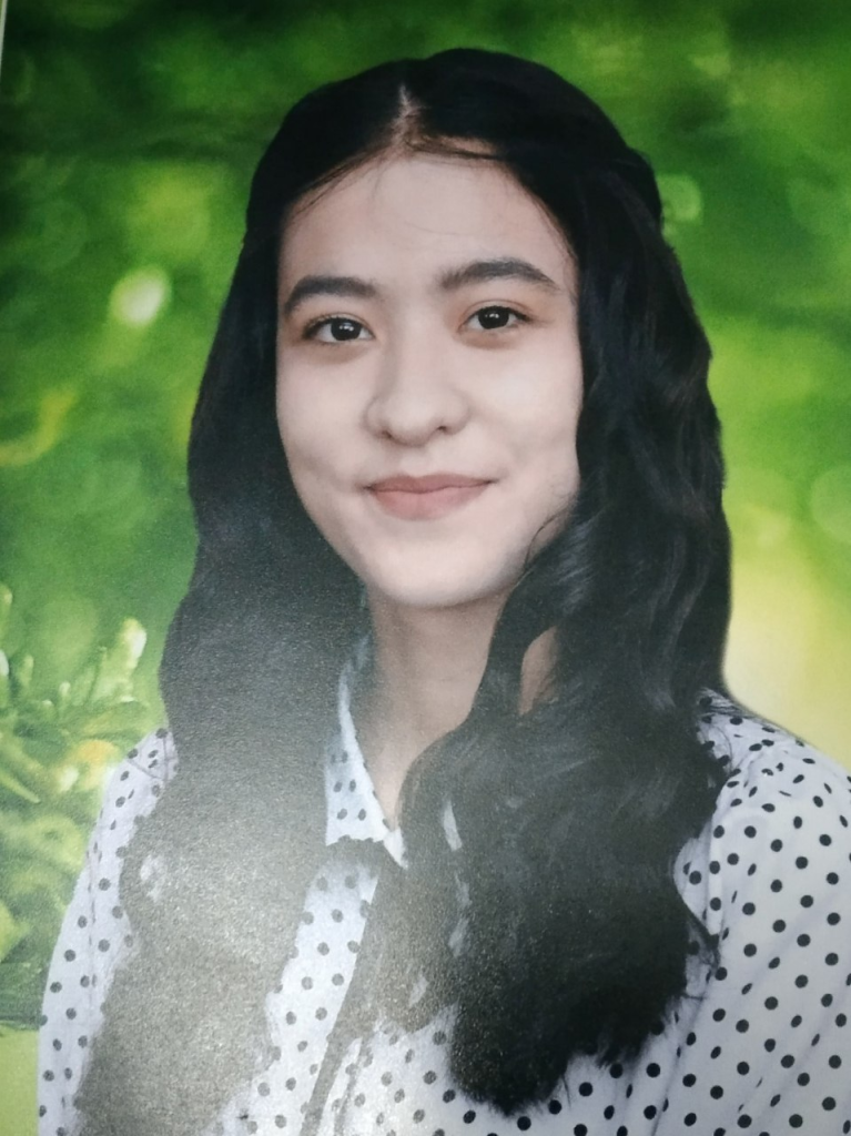 Young Central Asian woman with long slightly curly hair, black eyes, and a white blouse with black dots. She's got a green background behind her. 
