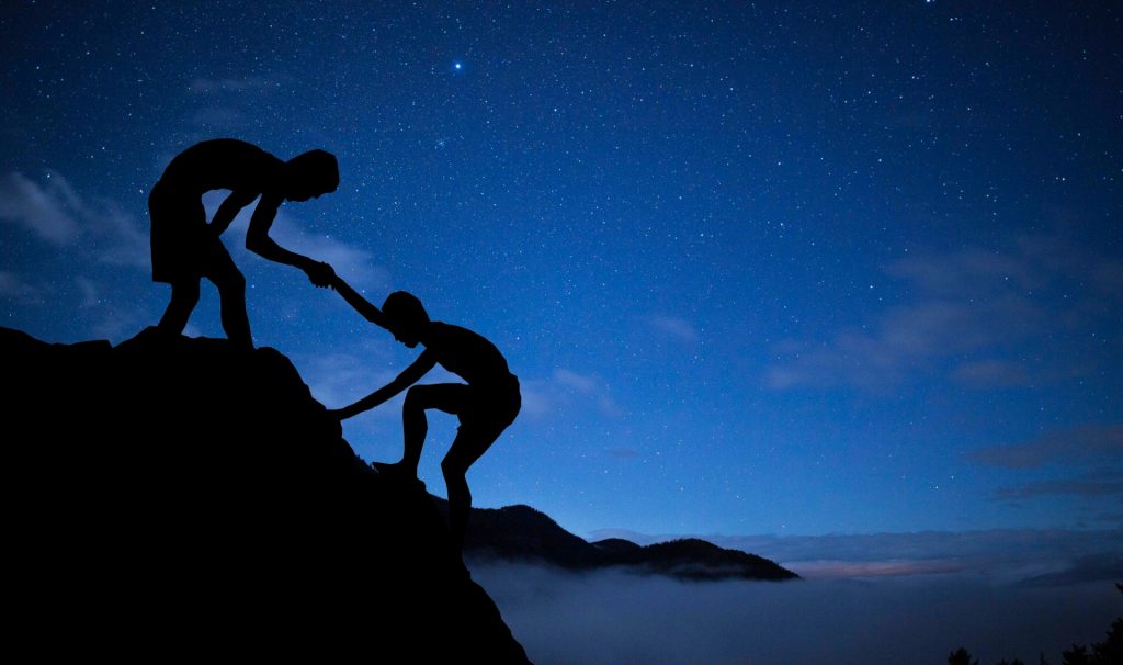 Rock climbers out on a starry night above some misty fog. One person bends to help another up a cliff. 