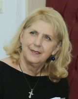 Older middle aged light skinned woman with earrings, light blonde hair, a necklace and a black blouse