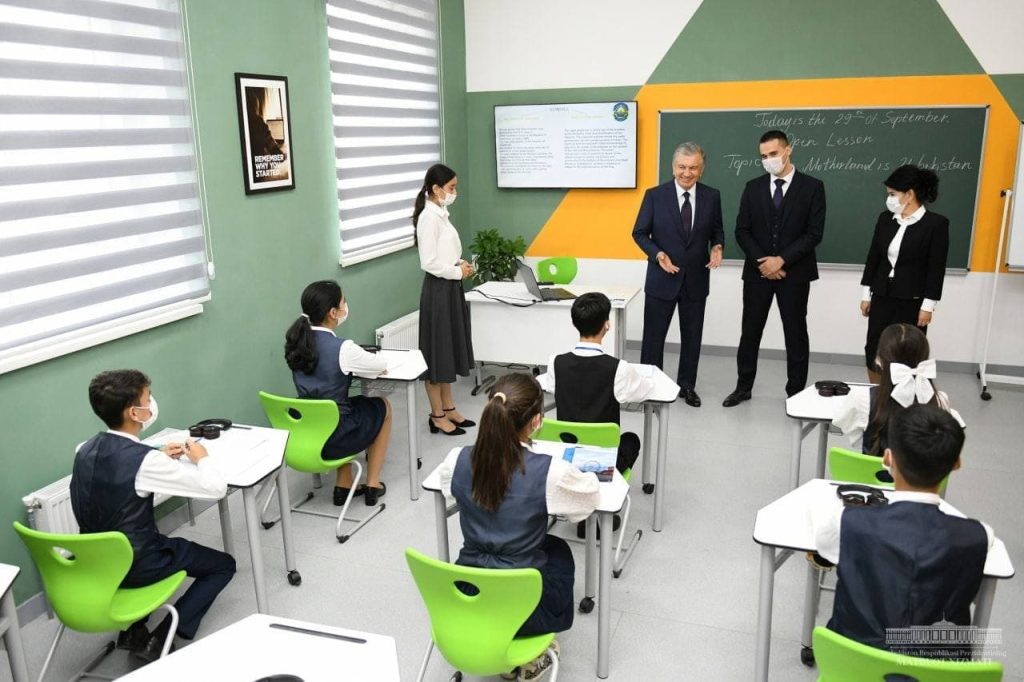 Gulsevar Xojamova, a young Central Asian student, stands up in the left front of a classroom that's green with white blinds, a green chalkboard, and a white floor. Three people in black, two men and a woman, are in front, and the students are in black and white uniforms and seated at white desks on green chairs. The author, Gulsevar, stands up near the front of the room. 