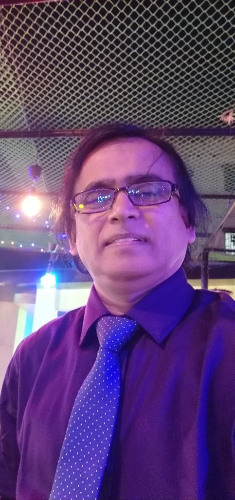Middle aged South Asian man with a purple suit and tie, reading glasses, and short brown shoulder length hair.