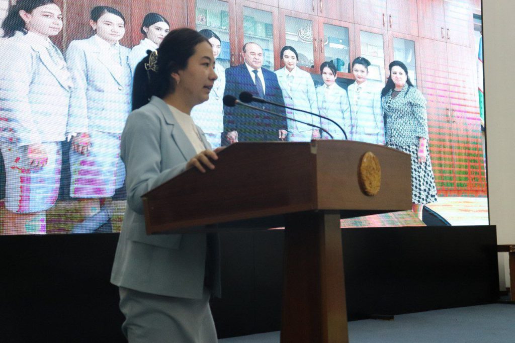 Central Asian woman with long dark hair in a ponytail and a light blue coat and dress pants speaks at a lectern. Behind her is a screen with several other similarly dressed women. 