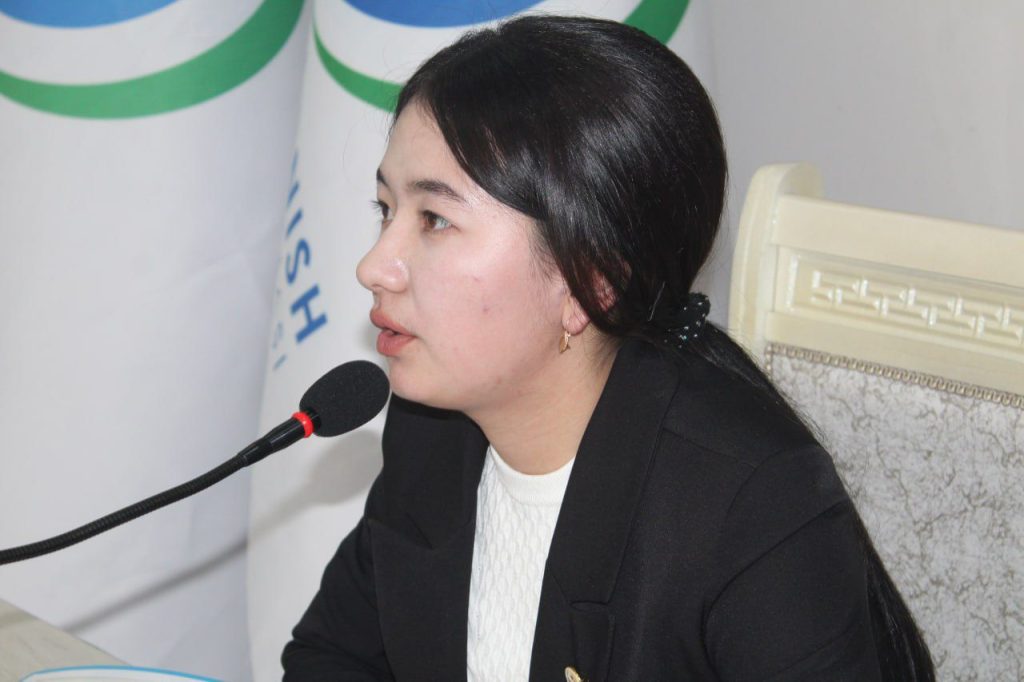 Young Central Asian woman with long dark hair and a black coat over a white blouse sits in front of a flag and microphone. 