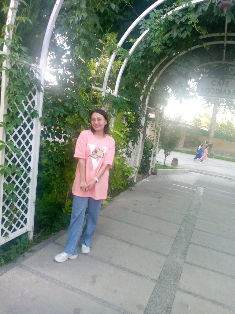 Young Central Asian woman with a pink tee shirt and jeans. She's standing in a passageway outside with green plants growing over a fence. 