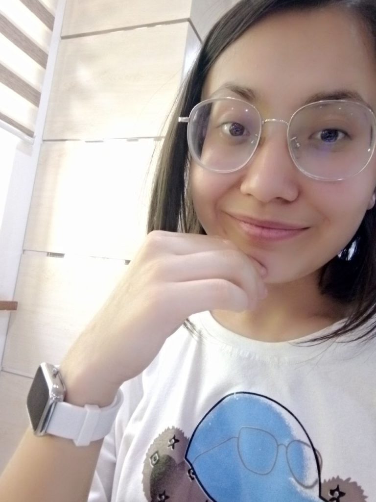Central Asian teen girl with reading glasses, short black hair, a white tee shirt with a blue design. She's got a wristwatch on her right hand which is near her mouth. 