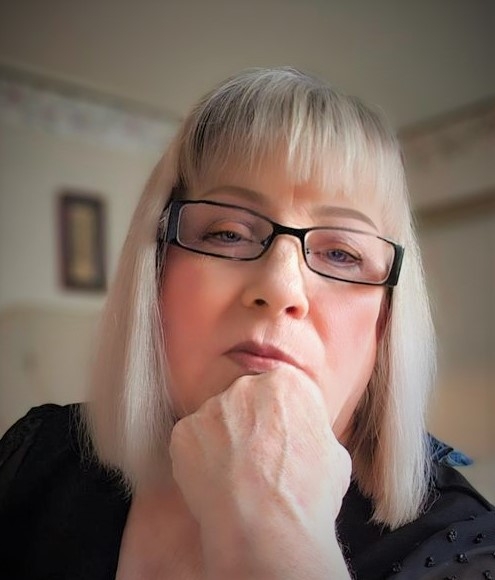 White middle aged woman with reading glasses and very blond straight hair resting her head on her hand.