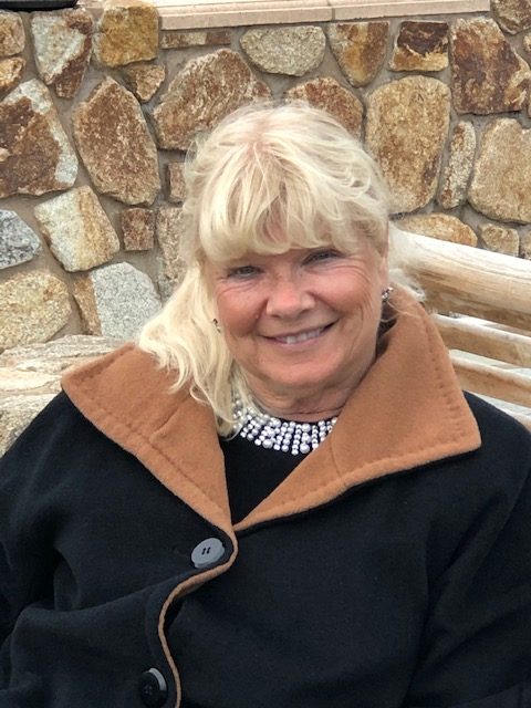 Older middle aged white woman with light blond hair and a large black jacket with a brown interior and a white beaded necklace. She's in front of a rock wall. 