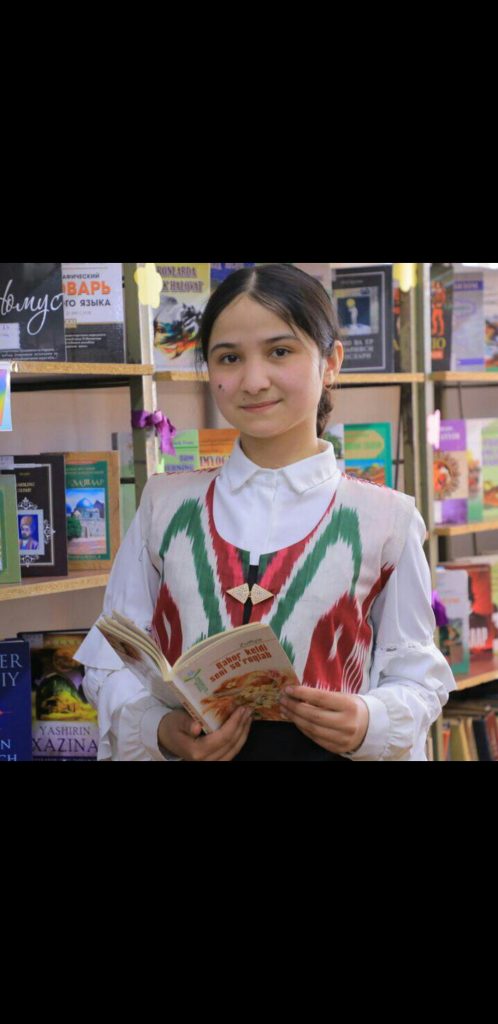 Central Asian teen girl with straight dark brown hair and a white outfit with red and green vertical stripes on top. She's standing in a classroom in front of a bookshelf. 