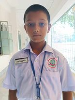 Young South Asian boy with short brown hair, white collared shirt with a school emblem and name tag, and a lanyard around his neck.